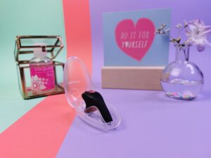 De fun factory be one ligt in de open bewaar case. Daar achter zie je een klein vaasje met een bloemetje, een kaart met de tekst 'do it for yourself' en daar naast staat een glazen kubus doosje met daarin een roze flesje sliquid.