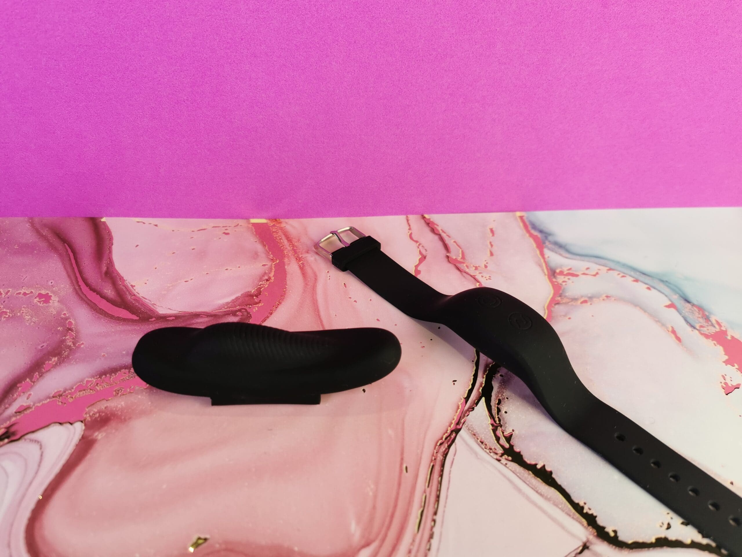 De CalExotics Lock-N-Play Wristband Remote Panty Teaser op een roze ondergrondje. De pantyvibrator is klein en zwart. Hij is langwerpig en heeft een hobbel voor de clitoris. Er zitten ribbeltjes over bijna de hele bovenkant. De afstandsbediening ziet er uit als een zwart smal horloge. Iets wat je bijvoorbeeld zou kunnen gebruiken om je stappen te tellen.