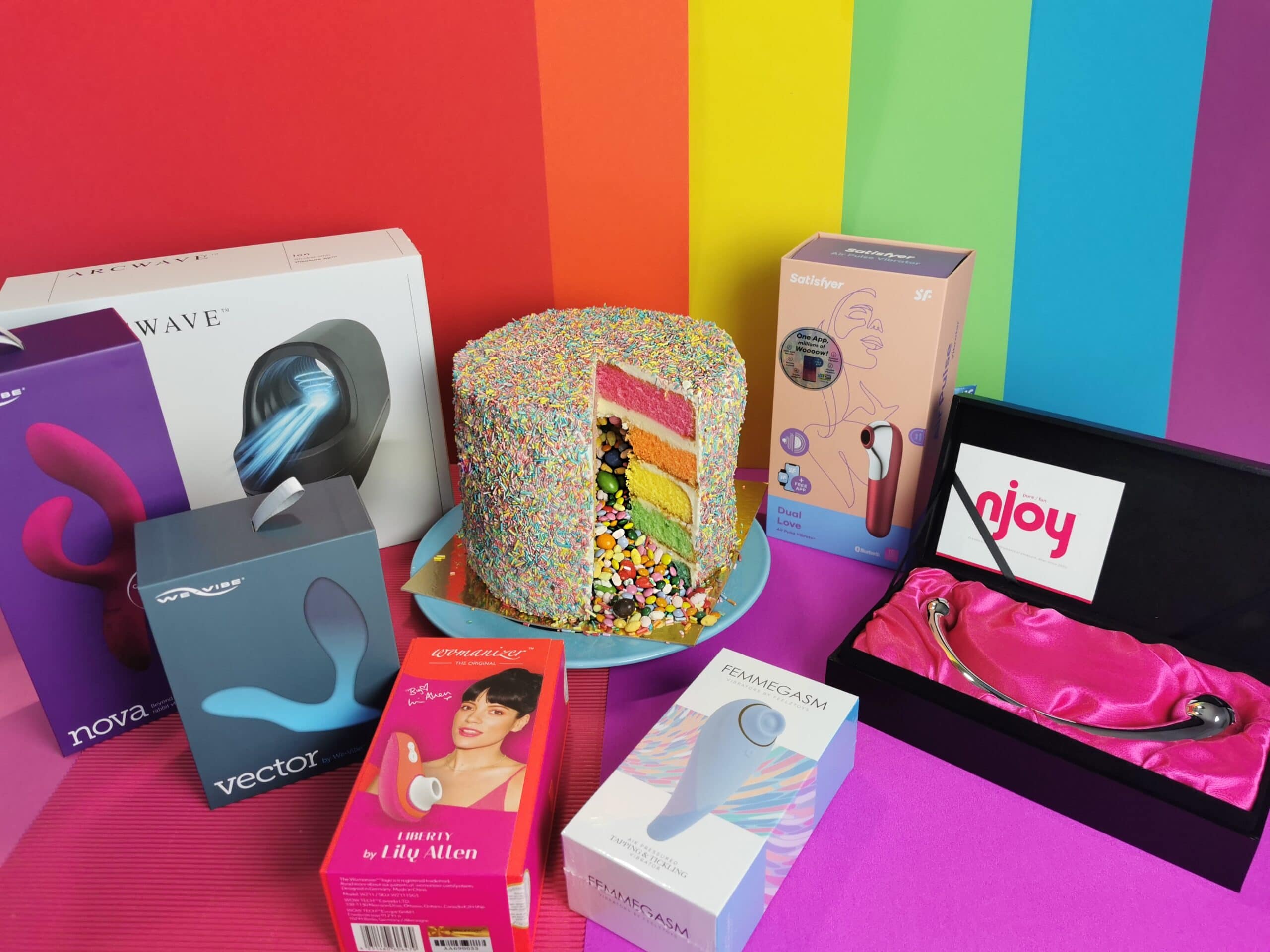 Op een roze en regenboog ondergrondje staat een regenboogtaart met heel veel sprinkles en snoepjes in het midden. Daar omheen staan alle toys die je kan winnen.