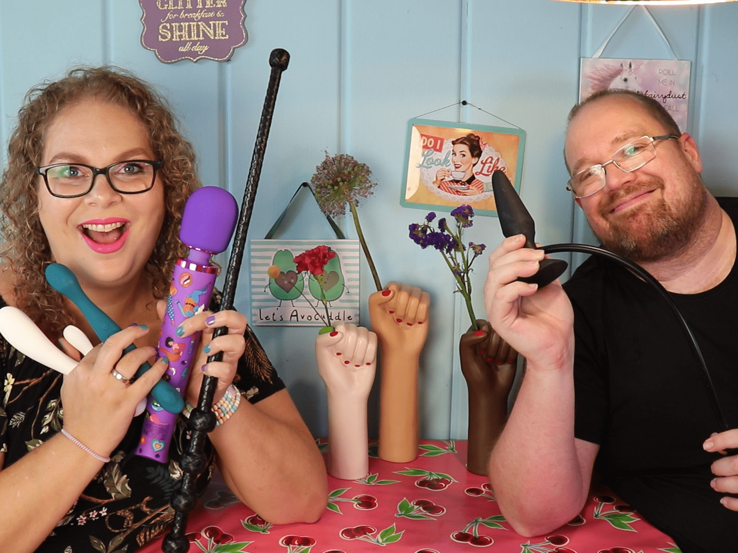 Tess en Jay zitten aan de keukentafel. Tess houdt een aantal sextoys vast en Jay houdt een opblaasbare buttplug vast.