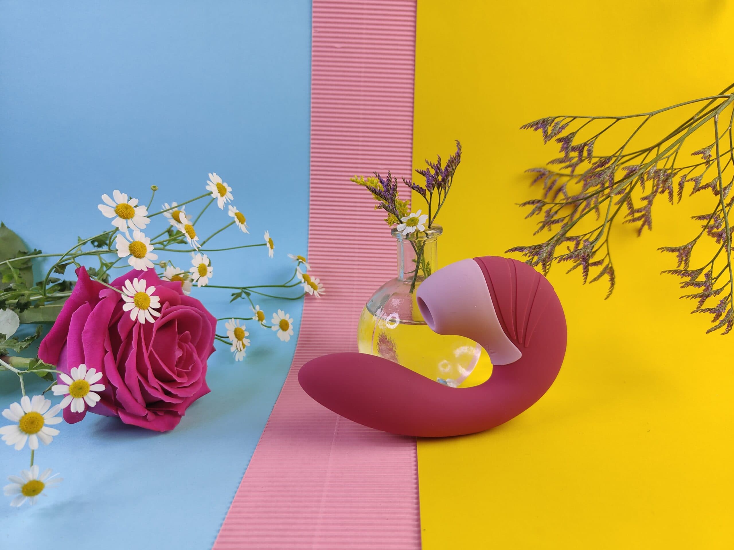 De Satisfyer Supernova op een blauw, roze en geel achtergrondje, met bloemen er omheen.