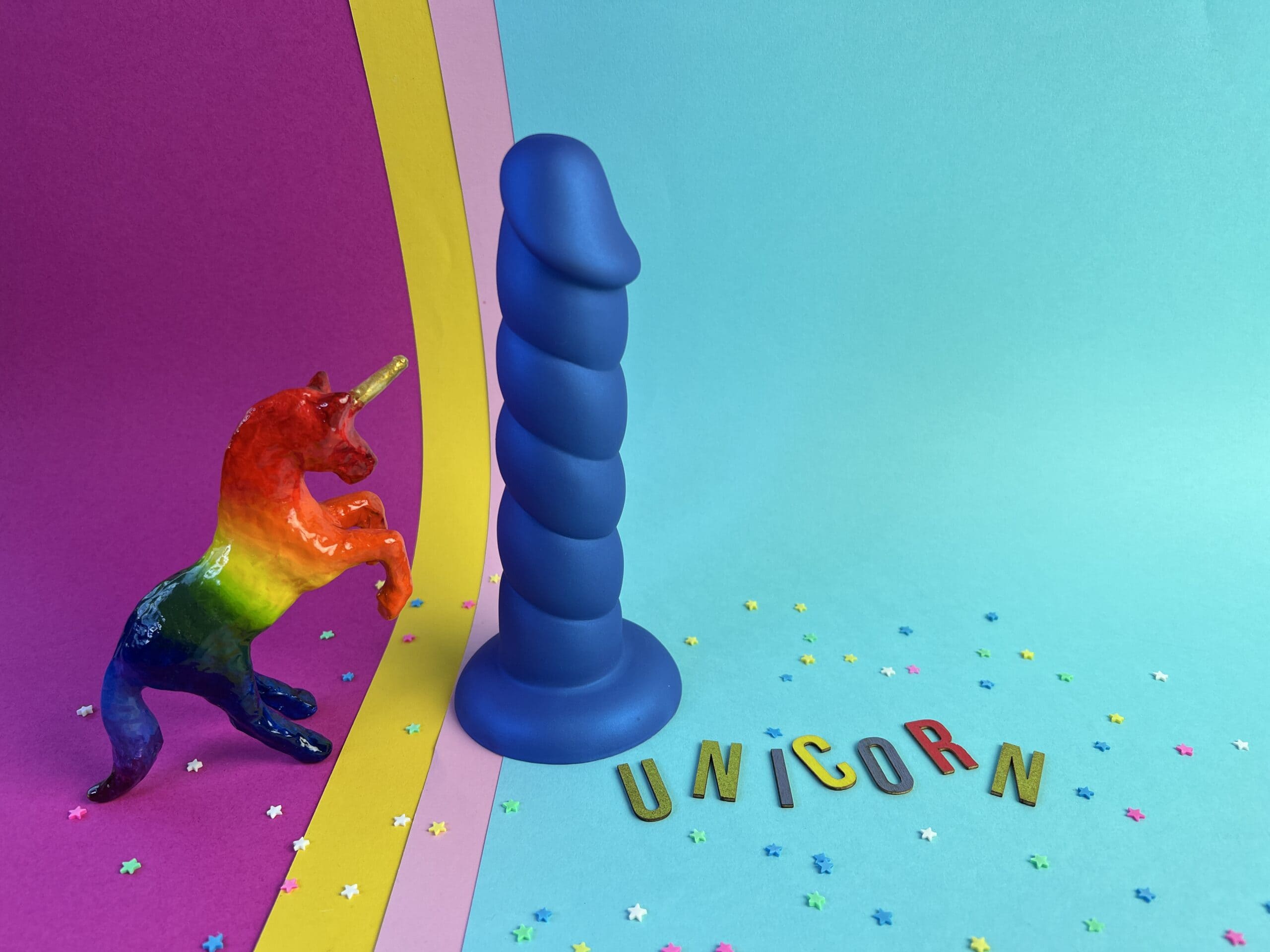 De Addiction Fantasy Unicorn Dildo naast een regenboog unicorn en er naast liggen de letters UNICORN