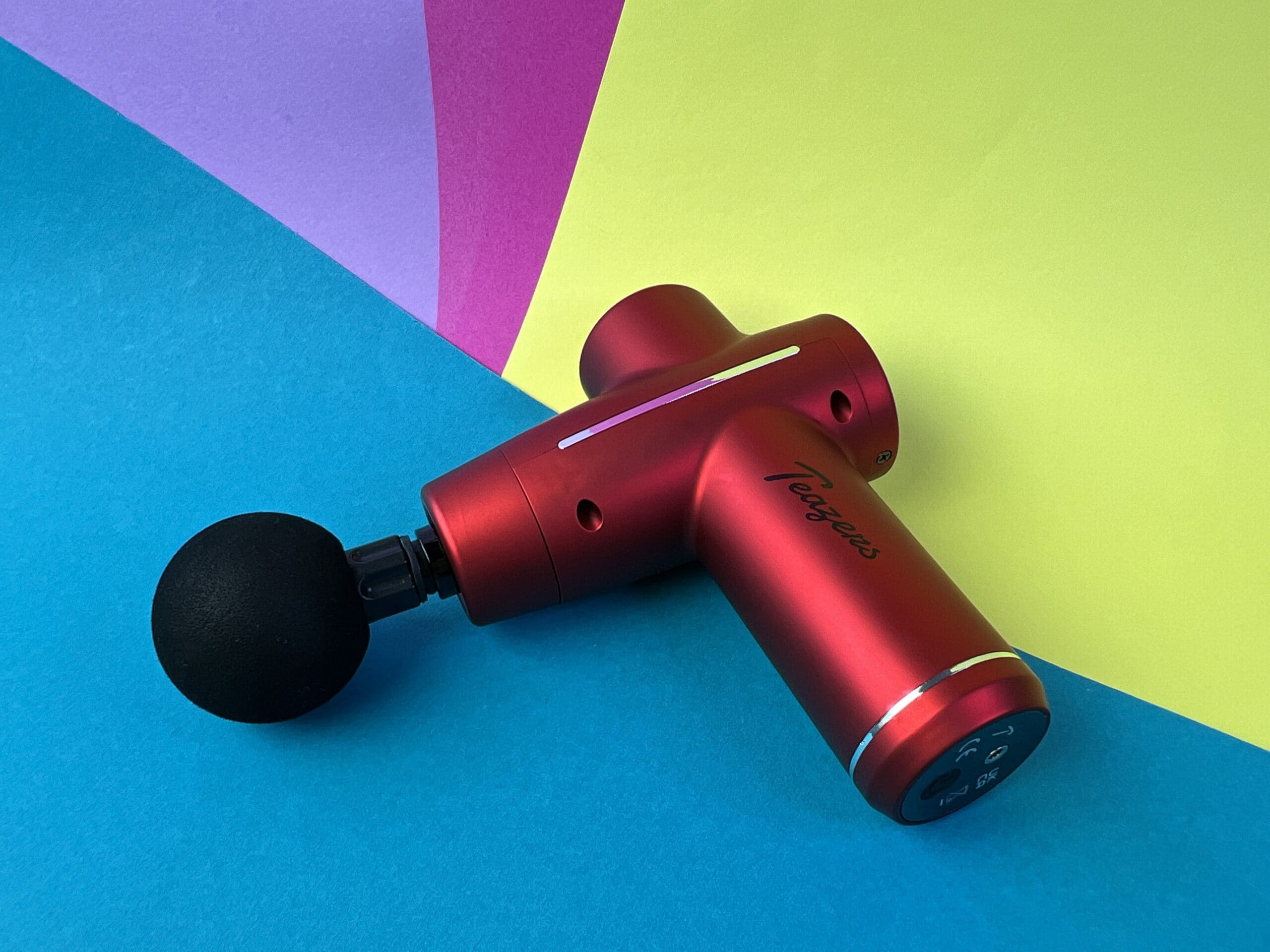 De Teazers Massage Gun op een meerkleurig ondergrondje.