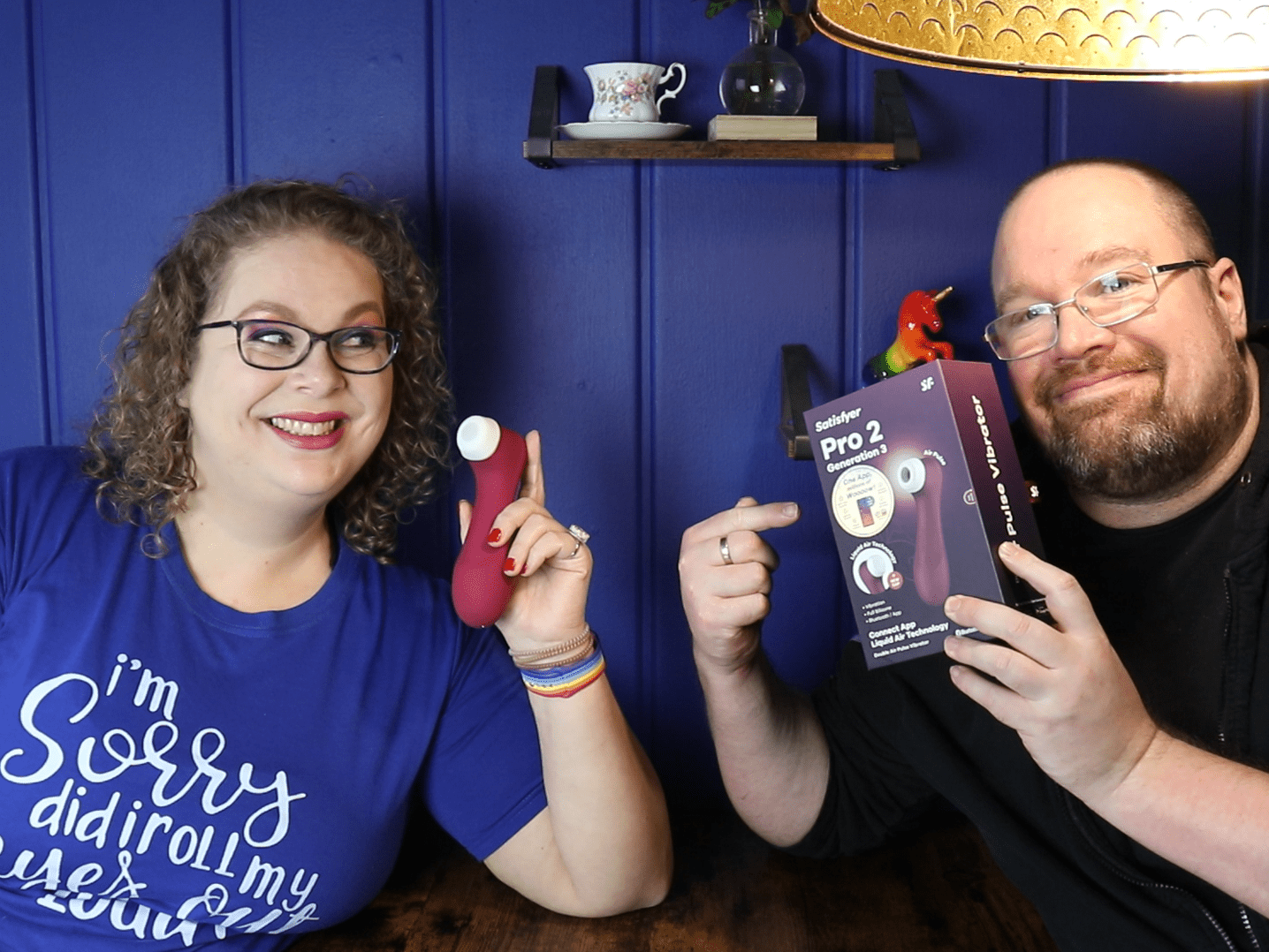 Tess en Jay zitten aan de keukentafel. Tess houdt de Satisfyer Pro 2 Generation 3 vast en Jay houdt de verpakking vast.