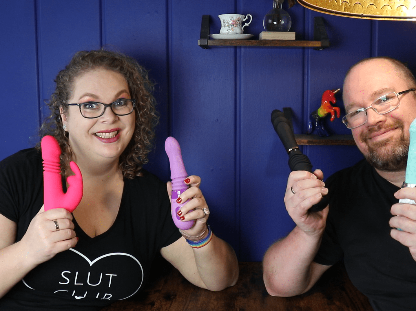 Tess en Jay zitten voor een donkerblauwe muur en houden allebei wat stotende sextoys vast.
