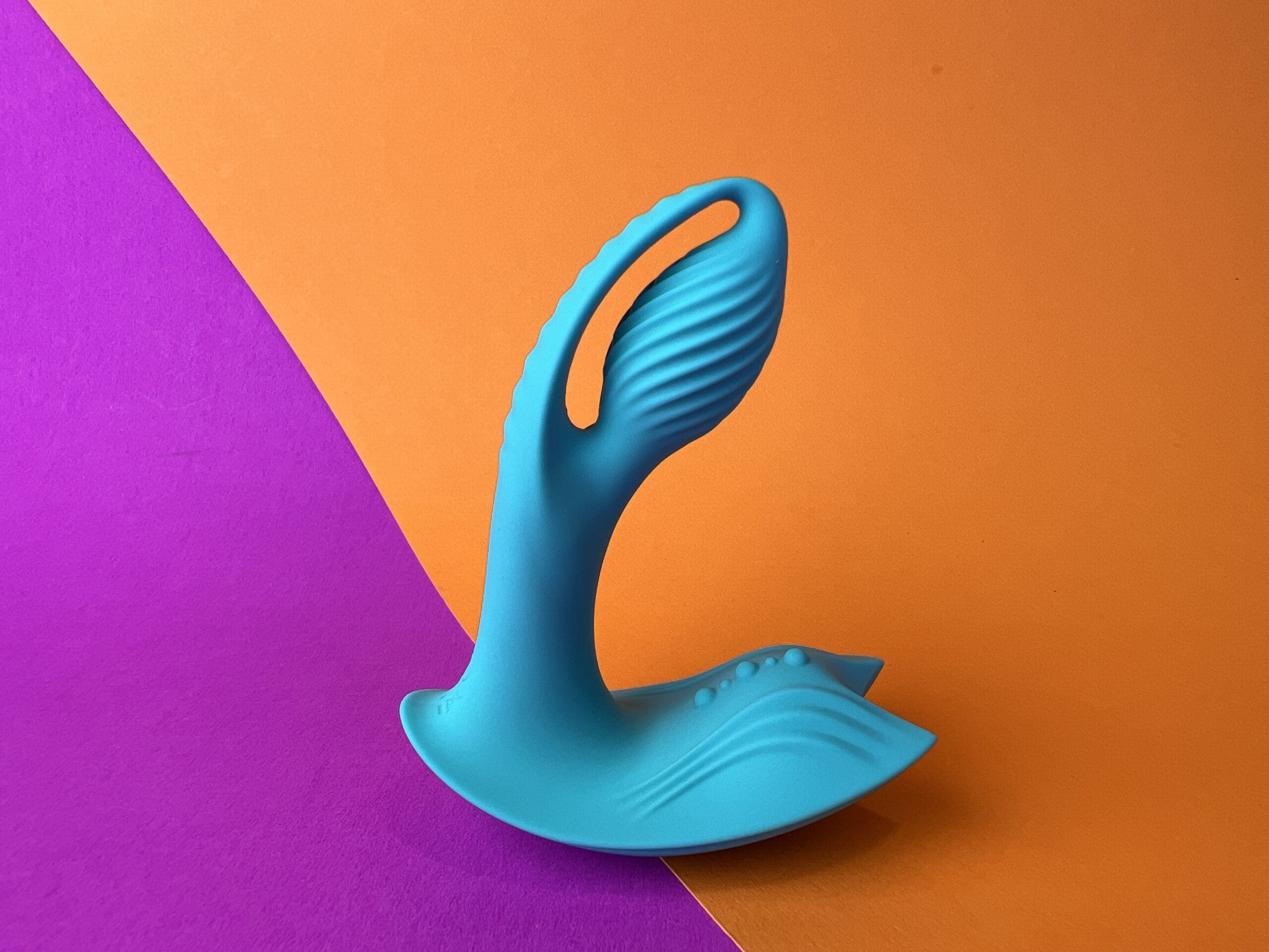 De Inya Bump-n-Grind is een mintgroene toy die wat op een prostaatstimulator of buttplug lijkt. Hij ligt op een oranje en roze ondergrondje.