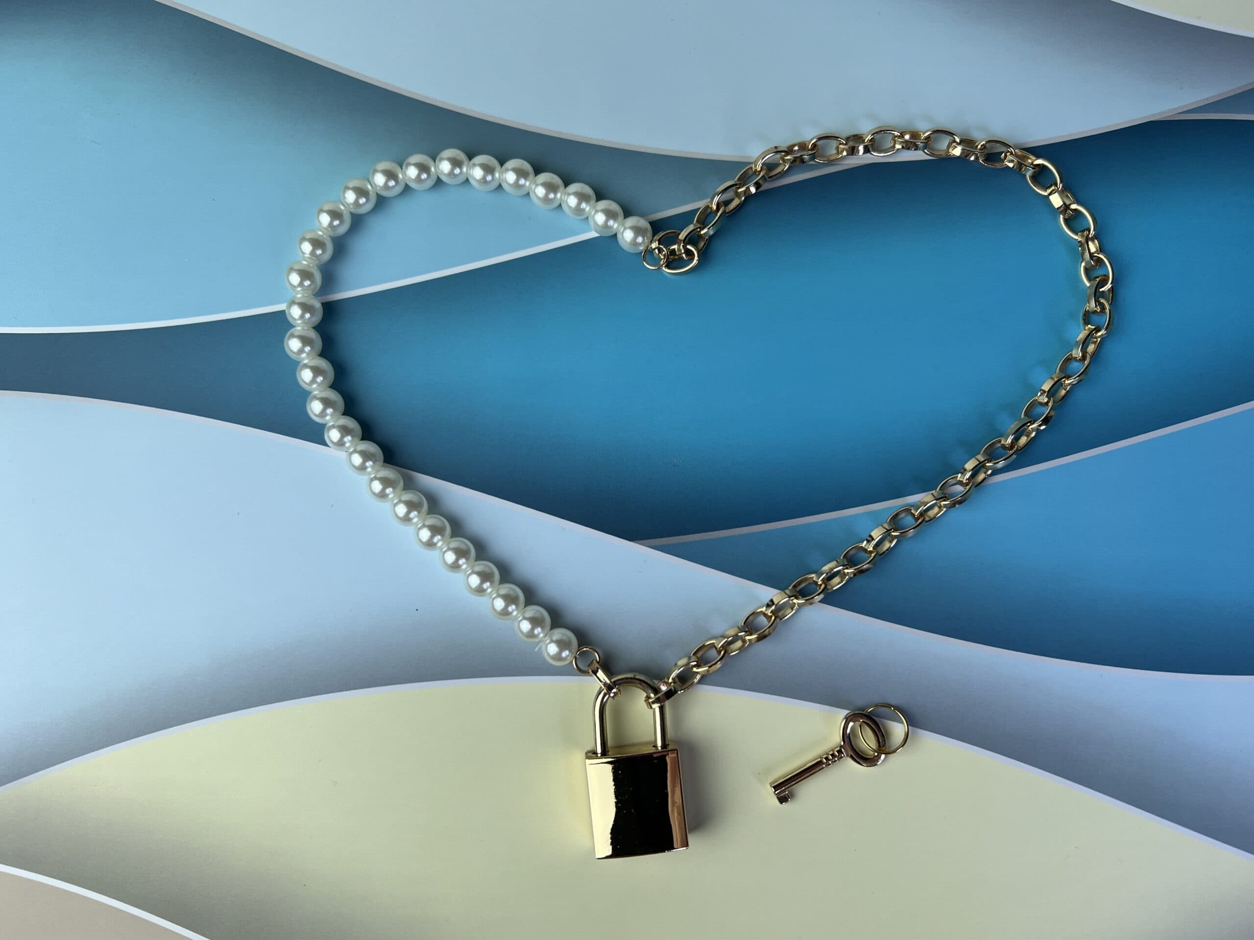 De Sex & Mischief Pearl Day Collar; de ene helft is een parelketting, de andere helft gouden ketting. De helften worden bij elkaar gehouden met een gouden hangslotje. Het sleuteltje ligt naast het slotje. De ketting ligt in een hart-vorm.