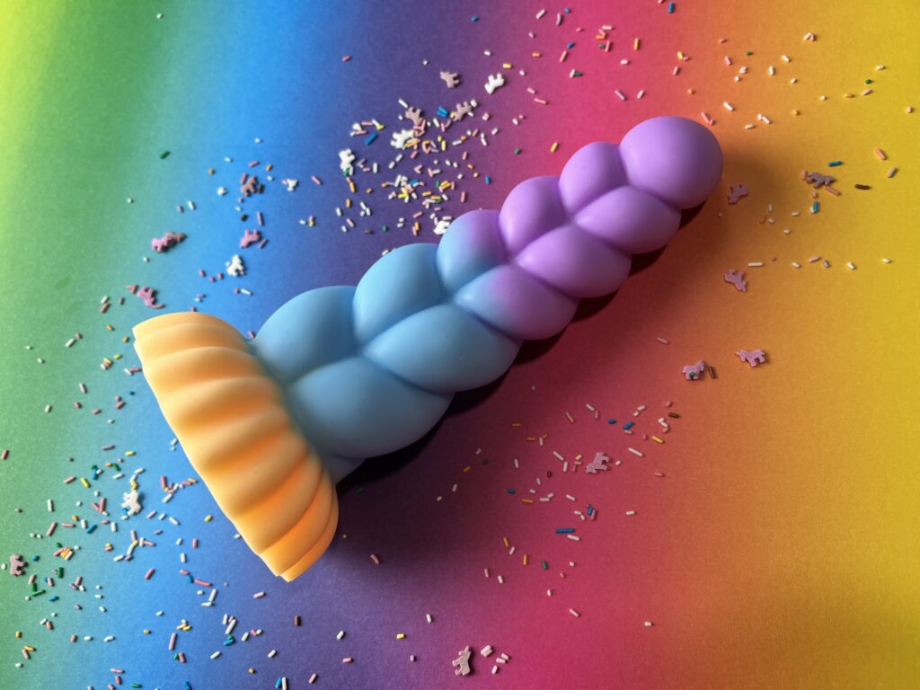 De creature cocks mystique unicorn dildo ziet er uit als de hoorn van een unicorn, maar dan dikker. De kleur verloopt van blauw naar paars en de onderkant is geel.