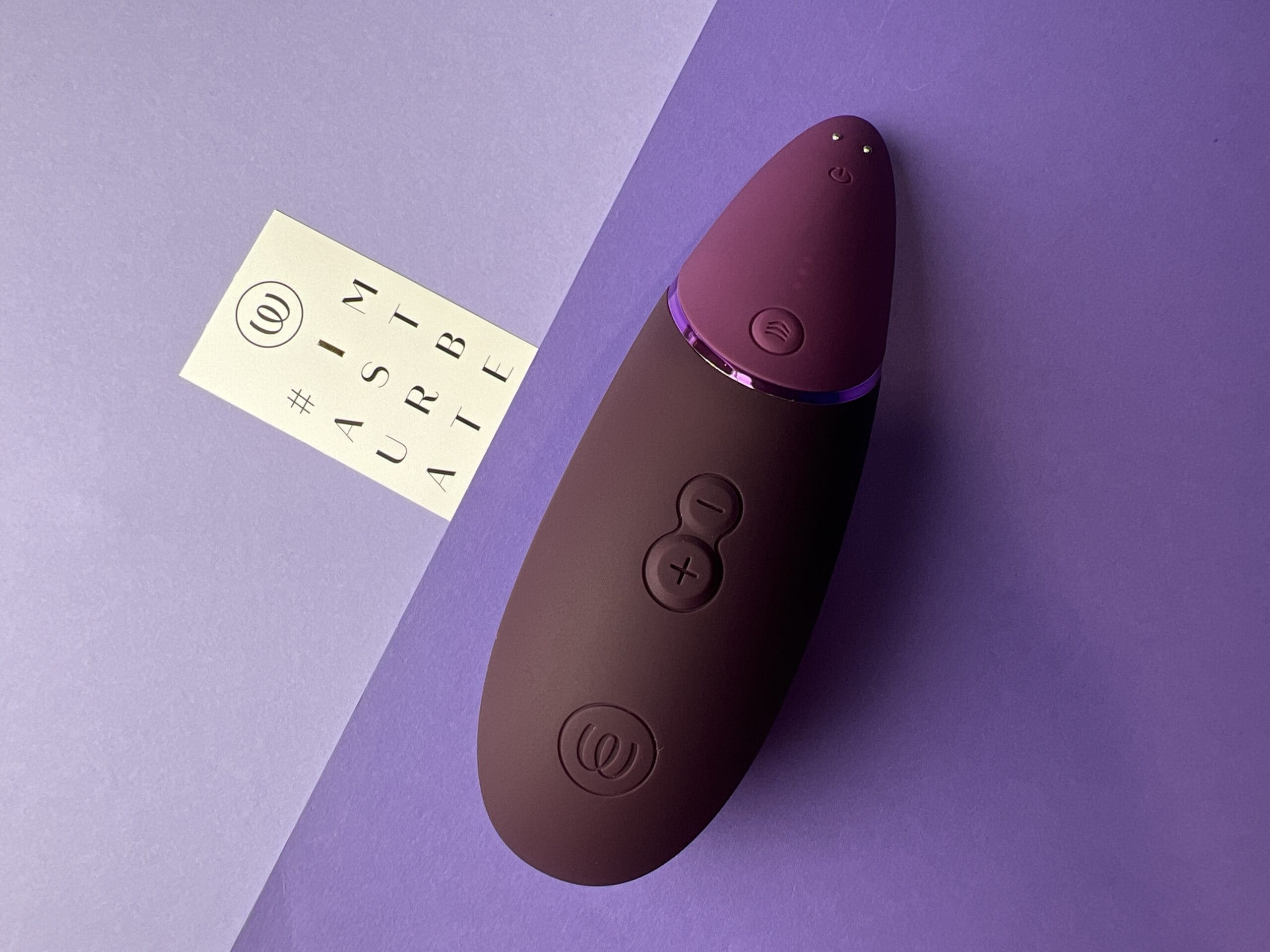 De Womanizer Next is twee tinten paars. Hij ligt ook op een ondergrondje in twee tinten paars met daarnaast een kaartje waarop staat I Masturbate.