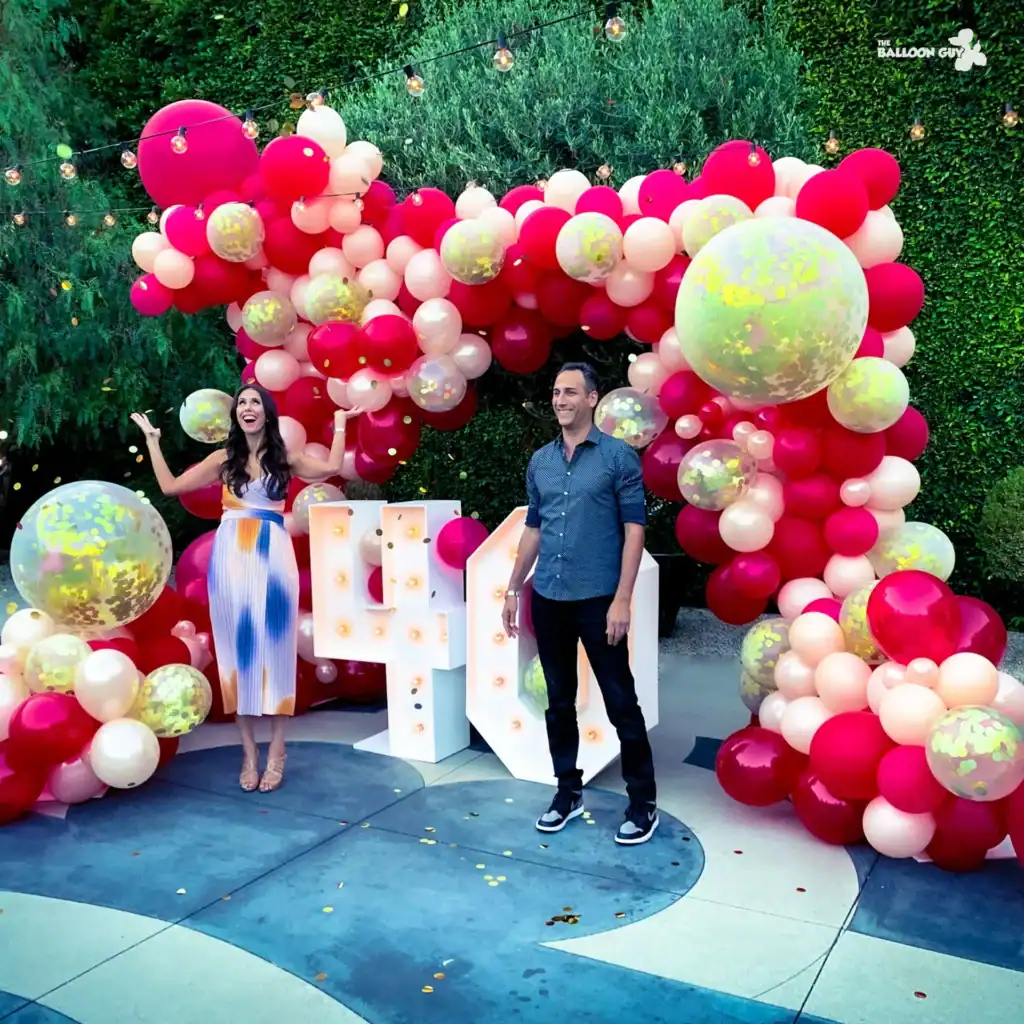 Two people stand in front of a stunning balloon arch crafted by a talented Los Angeles balloon artist, with red, pink, and white balloons accented by large light-up letters and outdoor string lights in the background.