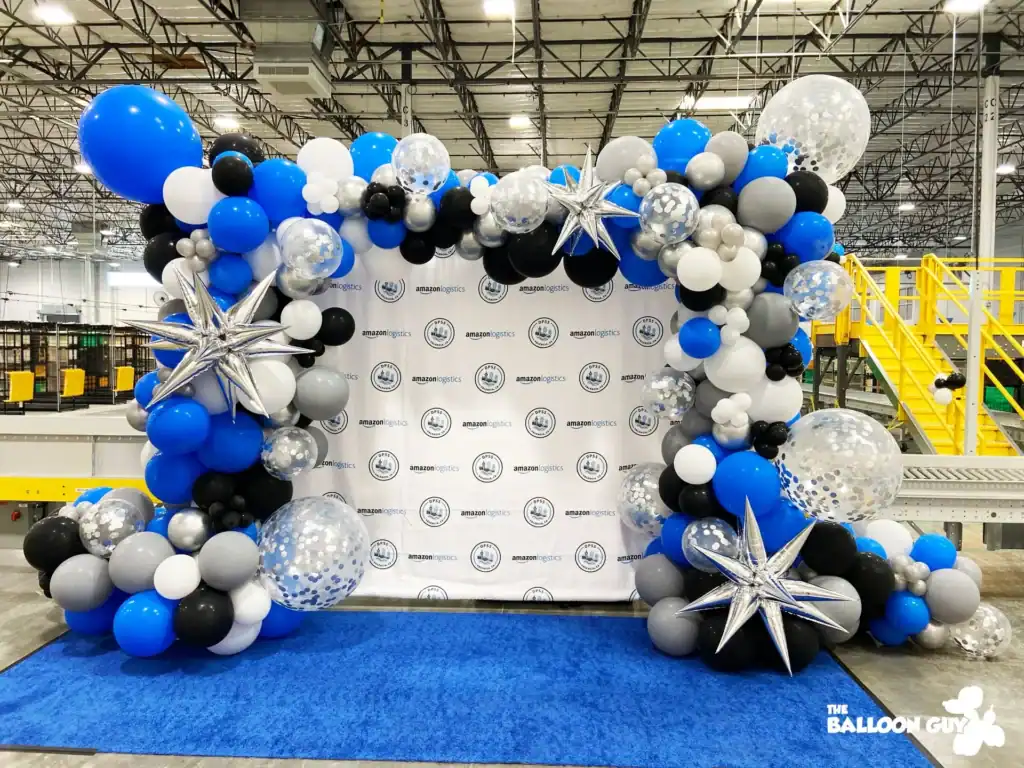 A stunning balloon arch with blue, black, white, and silver balloon decor and star accents frames a branded Amazon backdrop on a blue carpet—perfect for a corporate event in an industrial warehouse setting.