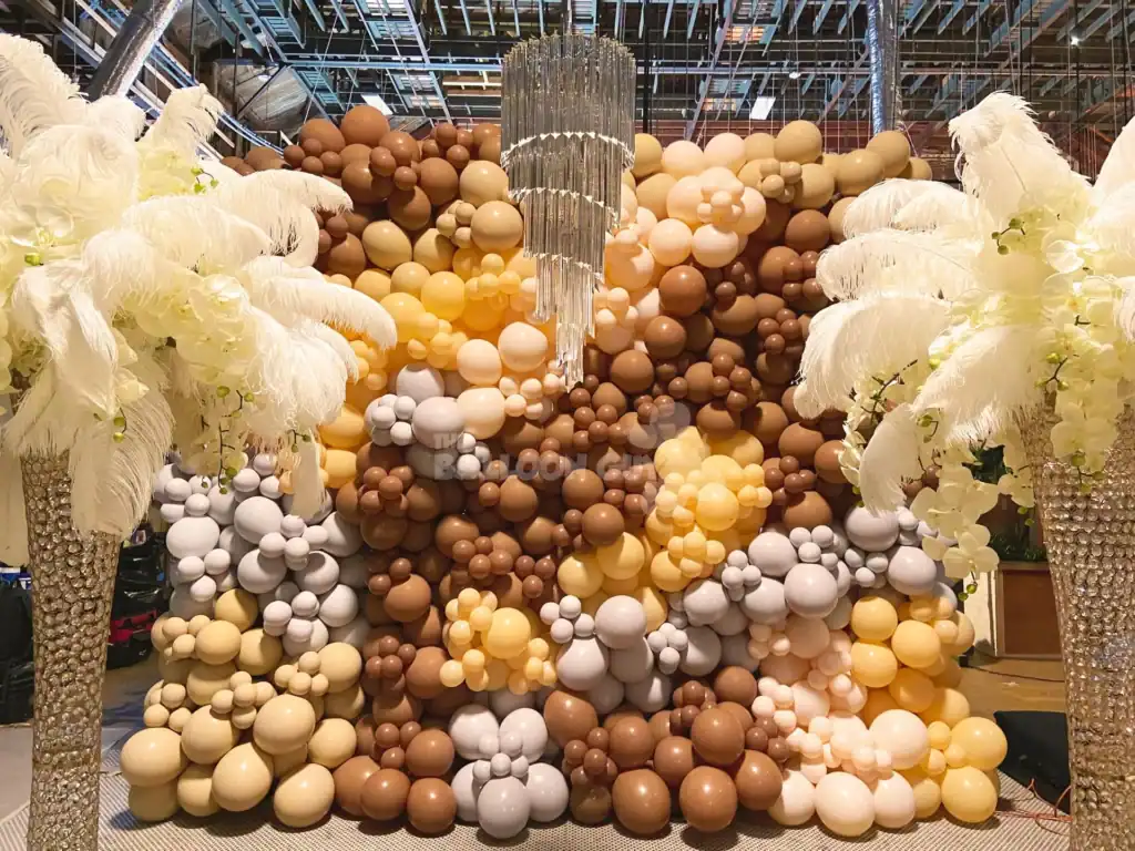 A decorative balloon wall in shades of brown, yellow, and white—crafted by a skilled balloon artist—stands under a chandelier, flanked by tall feathered arrangements on each side.