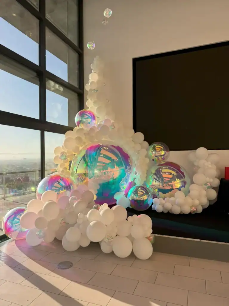 White and iridescent balloons of various sizes are arranged in a stunning balloon installation near large windows and a blank black screen in a modern indoor space.