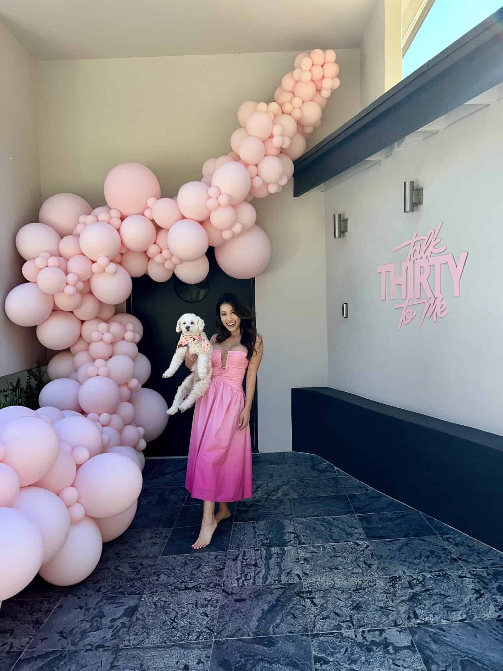 woman hold cute white puppy in front of house with pink balloon garland in los angeles by the balloon guy