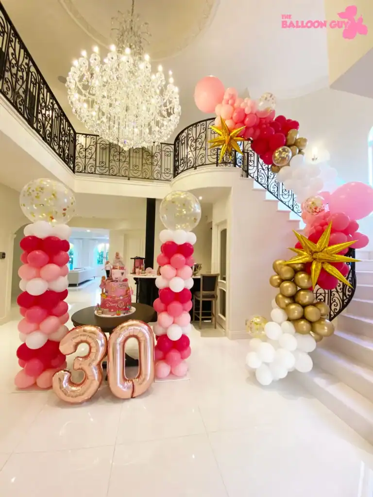 A stunning staircase and chandelier are adorned with balloon decor in pink, white, and gold, featuring a “30” balloon display and birthday cake at the heart of this stylish Los Angeles celebration setup.