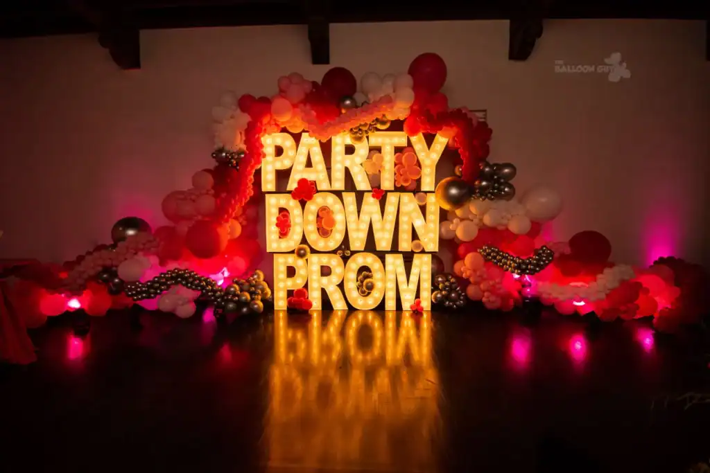 Large illuminated letters spell “PARTY DOWN PROM,” surrounded by vibrant balloon decor in red, white, pink, and silver, creating a festive atmosphere perfect for any corporate event. The dark floor and wall add dramatic contrast to the scene.