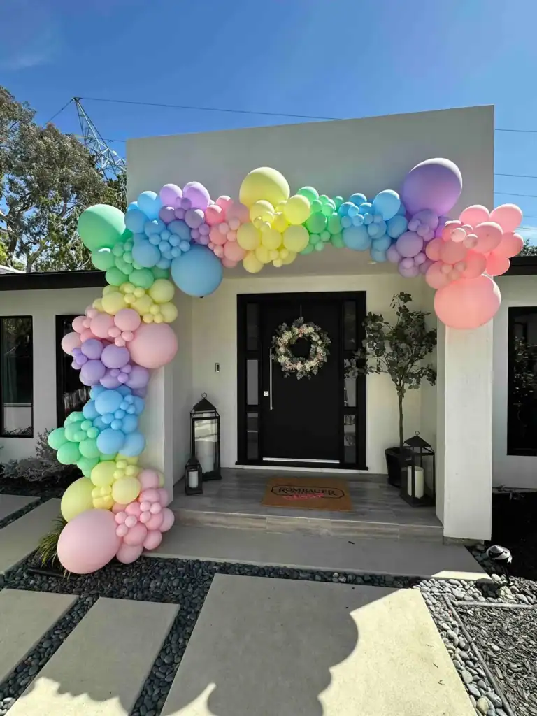 A pastel-colored balloon arch decorates the entrance of a modern Los Angeles house with a black front door and a wreath, flanked by large lanterns.
