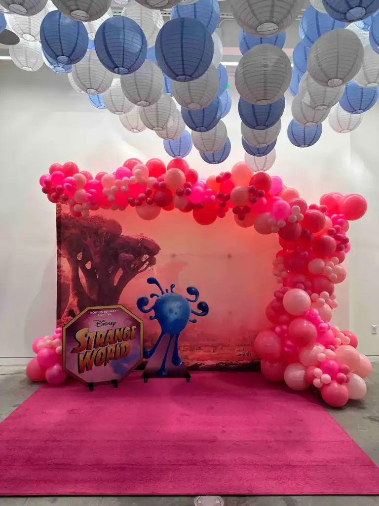 A decorated photo booth in Los Angeles with a Disney Strange World backdrop, pink and red balloons by a talented balloon artist, a blue character cutout, and blue paper lanterns hanging from the ceiling.
