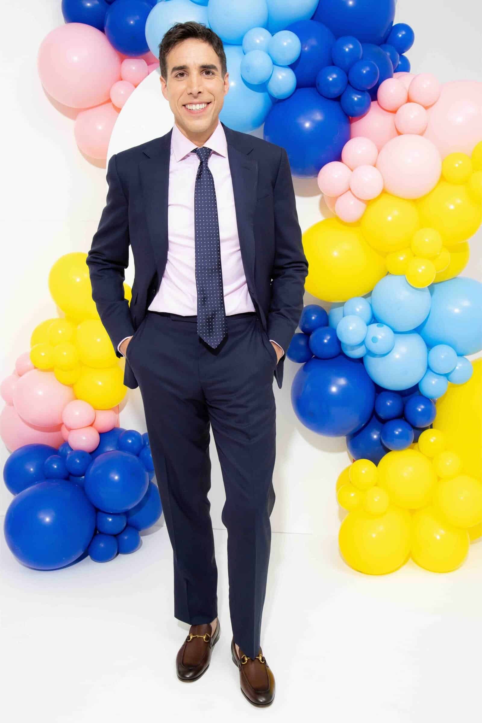 A man in a navy suit and tie stands smiling in front of a vibrant balloon installation featuring blue, yellow, and pink balloons.