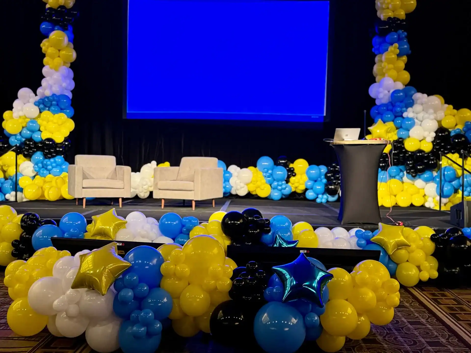 Stage in Los Angeles decorated with stunning balloon installations in blue, yellow, black, and white, two beige armchairs, a podium with a laptop, and a large blank blue screen in the background.
