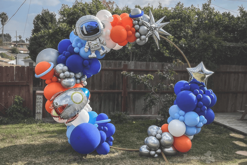 An arch made of blue, orange, silver, and white balloons with space-themed decorations—planets, stars, and an astronaut—set up outdoors on grass. Perfect balloon decor for a corporate event or memorable celebration.
