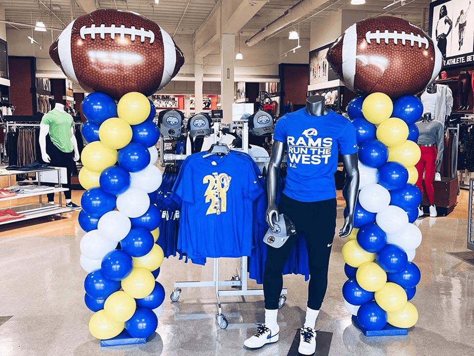 A store display features striking balloon installations with football balloon columns, blue and gold shirts on racks, and a mannequin dressed in “Rams Run the West” apparel.
