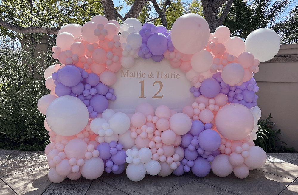 A pastel balloon display by a talented balloon artist in Los Angeles features pink, purple, and white balloons surrounding a sign reading "Mattie & Hattie 12" outdoors.