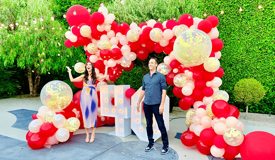Two people stand smiling in front of a vibrant balloon decor arch with large light-up numbers, outdoors with greenery in the background.