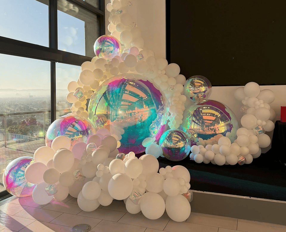 A large decorative balloon display, crafted by a skilled balloon artist, features clusters of white and iridescent balloons set up indoors near large windows and a stage area—perfect for a stylish Los Angeles corporate event.
