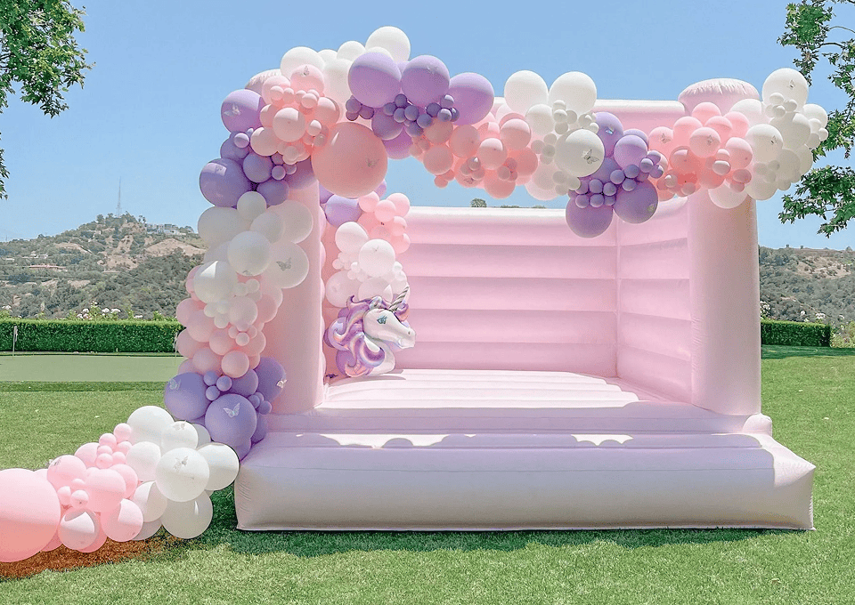 A pastel pink inflatable bounce house decorated with balloon decor in shades of pink, white, and purple is set up outdoors on grass, with trees and hills in the background.