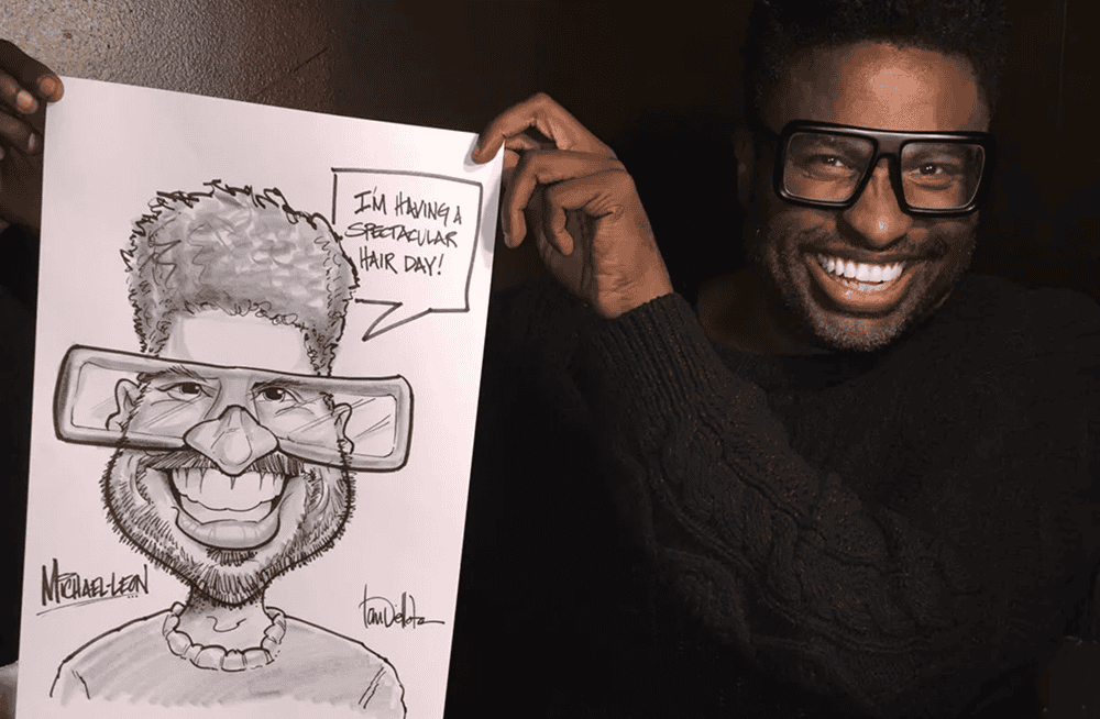 A man smiles while holding a caricature of himself, featuring exaggerated glasses and hair, with a sign that reads, “I’m having a spectacular hair day!” at a balloon decor event in Los Angeles.