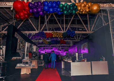 An indoor corporate event space in Los Angeles features a red carpet, colorful balloon artist decorations on the ceiling, and two people walking toward a purple-lit area.