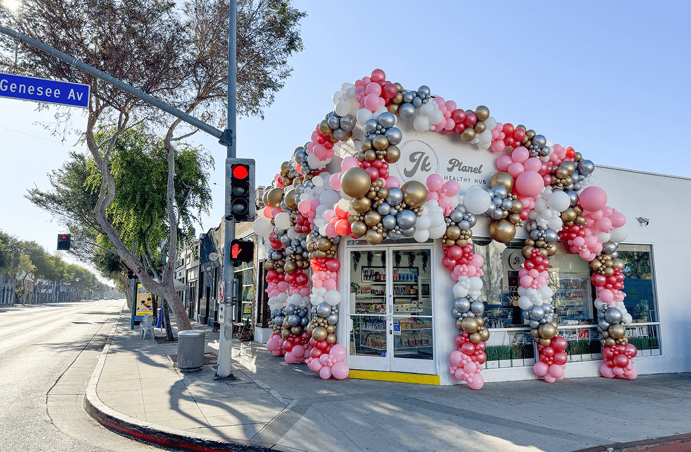 A white storefront in Los Angeles, decorated with pink, white, gold, and silver balloons by a talented balloon artist on the corner of Genesee Ave and a quiet street, with a traffic signal showing red. Perfect for hosting your next corporate event.