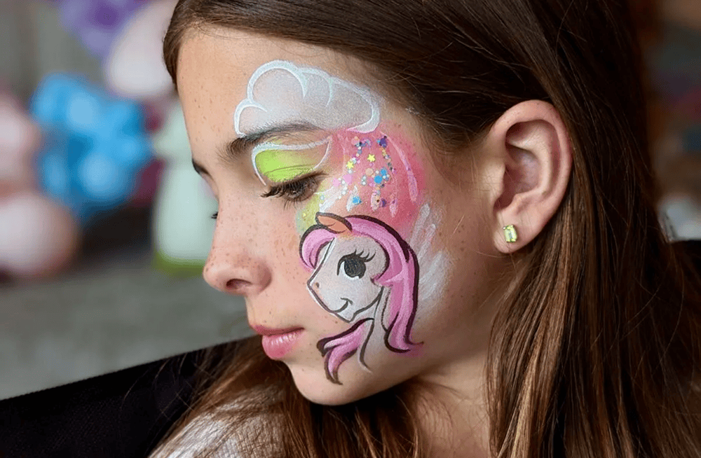 A girl with long brown hair has her face painted with a unicorn, clouds, and colorful stars on her cheek and around her eye at a vibrant Los Angeles corporate event with stunning balloon installations.