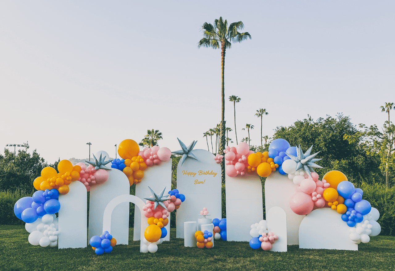 Outdoor birthday party setup in Los Angeles with white arches decorated with colorful balloons in blue, orange, pink, and purple; "Happy Birthday Sam!" written on the center arch. Palm trees sway gently in the background.
