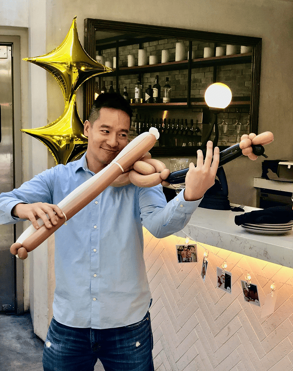 A man in a blue shirt pretends to play a violin made of balloons in a modern indoor setting, featuring creative balloon decor with two gold star balloons and photos hanging on the bar—perfect for a lively corporate event.