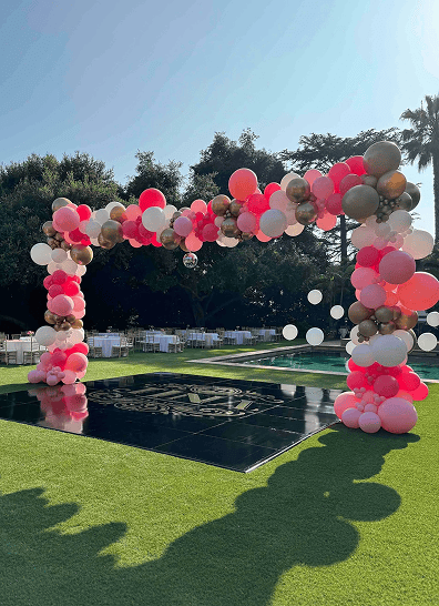 A large outdoor balloon arch created by a skilled balloon artist features pink, white, and gold balloons framing a black dance floor on a grassy lawn. Tables and trees add to the festive scene in the background.