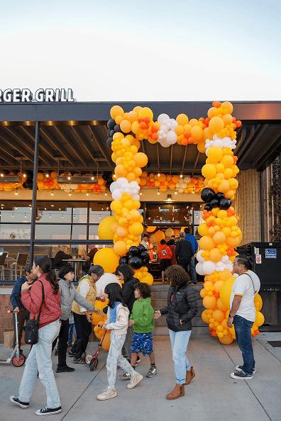 A group of people waits outside a restaurant decorated with vibrant balloon decor, featuring a large yellow, orange, white, and black balloon arch at the entrance.