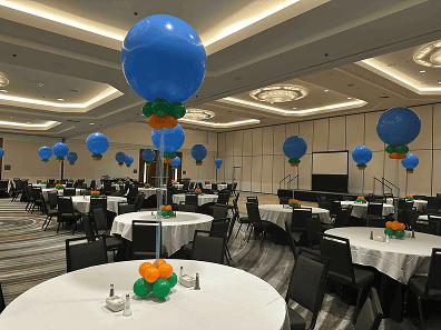 A banquet hall set for a corporate event features round tables with white tablecloths, each adorned with elegant balloon decor in blue and orange. Black chairs are arranged neatly around the tables.