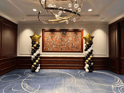 An empty room with dark wood paneling, blue carpet, a large abstract painting, and elegant balloon decor with gold star toppers stands under a modern chandelier—perfect for hosting your next corporate event.