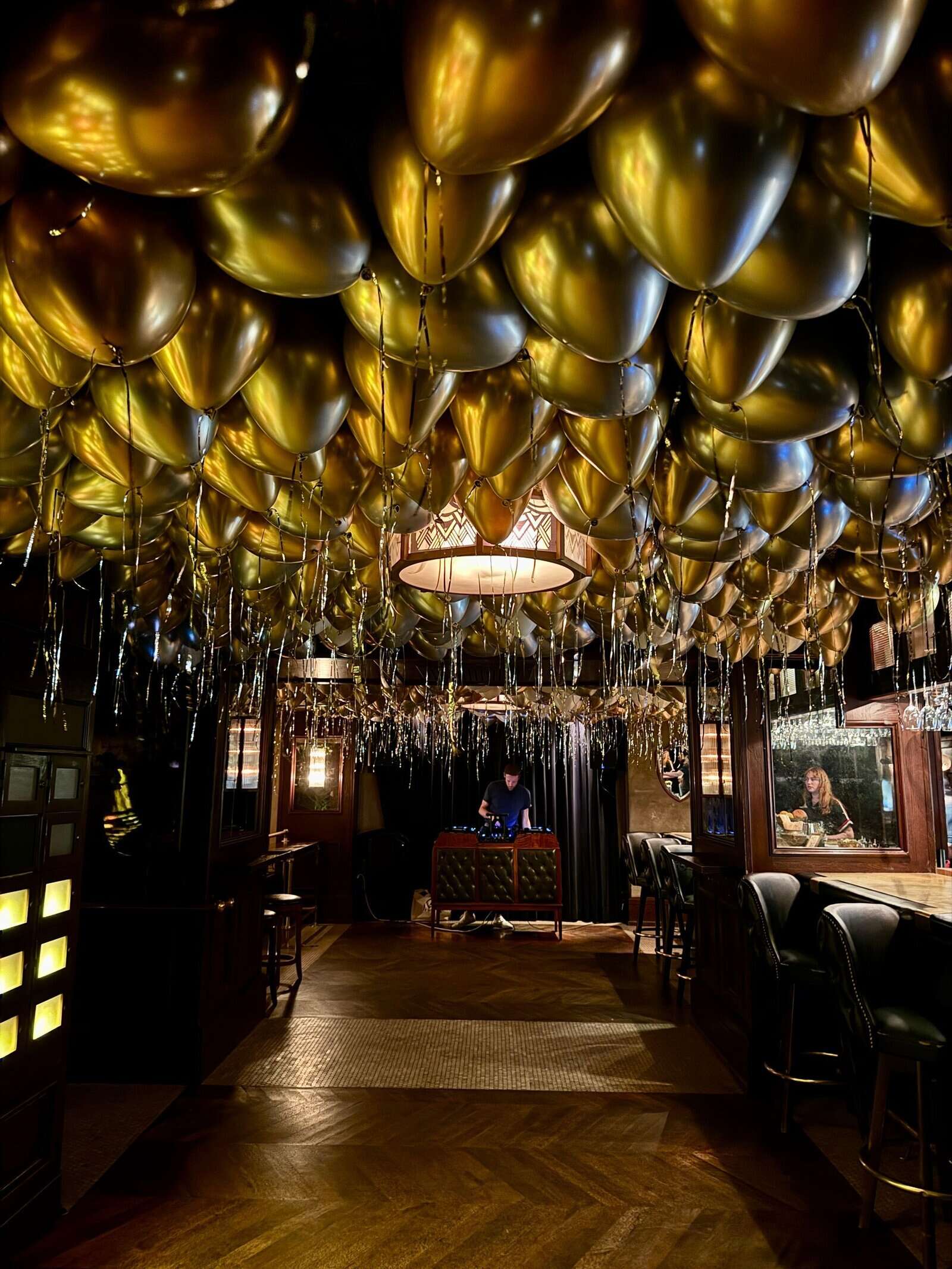 A dimly lit Los Angeles bar features striking balloon installations with gold balloons covering the ceiling and metallic streamers hanging down; one person sits at a table and another at the bar.