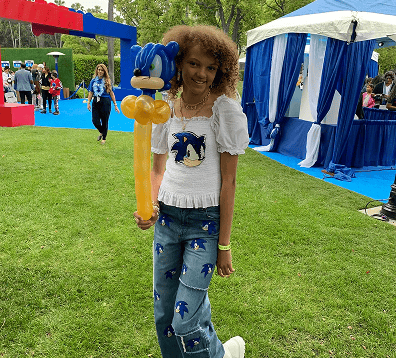 A person stands on grass holding a Sonic the Hedgehog balloon, wearing a white Sonic-themed shirt and blue jeans with Sonic graphics. Blue and white tents and creative balloon decor are in the background.