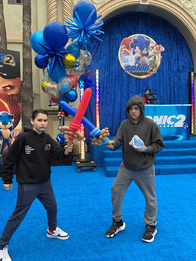 Two kids on a blue carpet hold balloon swords and balloons at a Sonic the Hedgehog 2 event, with colorful balloon installations and a Sonic backdrop decorating the space behind them.