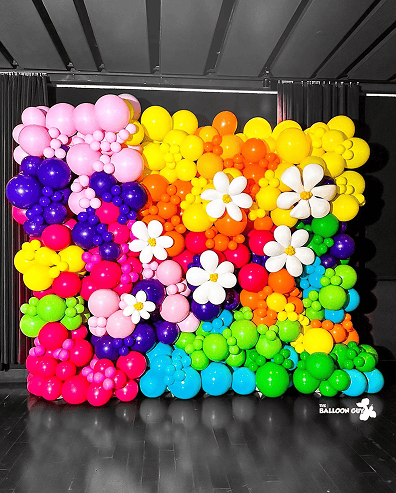 A colorful wall of balloons arranged in clusters and flower shapes, created by a talented balloon artist in Los Angeles, features bright shades of pink, yellow, orange, purple, green, and blue against a dark background.