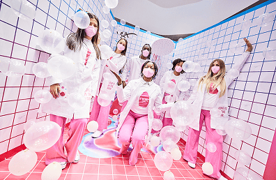 Six people in white lab coats and pink pants stand in a colorful Los Angeles room with grid walls and floating balloons, all wearing pink face masks.