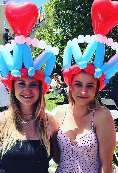 Two women outdoors in Los Angeles wear colorful balloon hats with red heart shapes on top, smiling at the camera. White chairs and greenery are visible in the background, showcasing the playful work of a talented balloon artist.