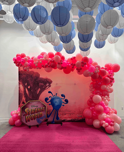 A pink balloon arch by a talented balloon artist frames a "Disney Strange World" sign and blue character, all set on a pink carpet; blue and white paper lanterns hang from the ceiling at this Los Angeles event.