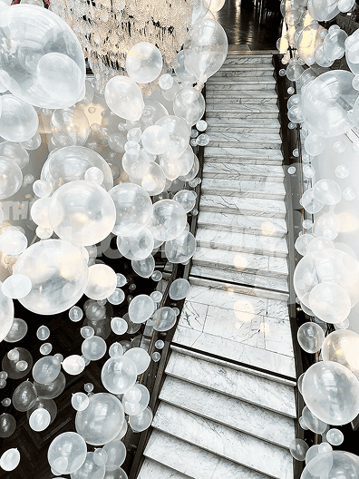 A marble staircase in Los Angeles is surrounded by numerous translucent white balloons of various sizes, creating a floating effect along the steps and banisters, masterfully arranged by a skilled balloon artist.