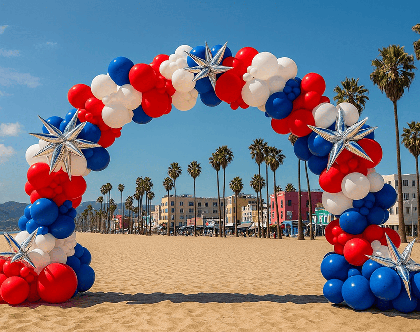 A red, white, and blue balloon arch with silver star decorations, crafted by a skilled balloon artist, stands on a sandy beach with palm trees and colorful buildings in the background.