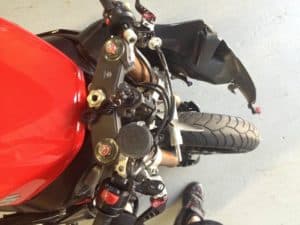 From above, a red motorcycle's front end reveals its handlebars, brake fluid reservoir, and front tire. Nearby, on a light gray surface, is Clay Green's foot in a black shoe—the perfect stance of a Flow & Performance Coach.